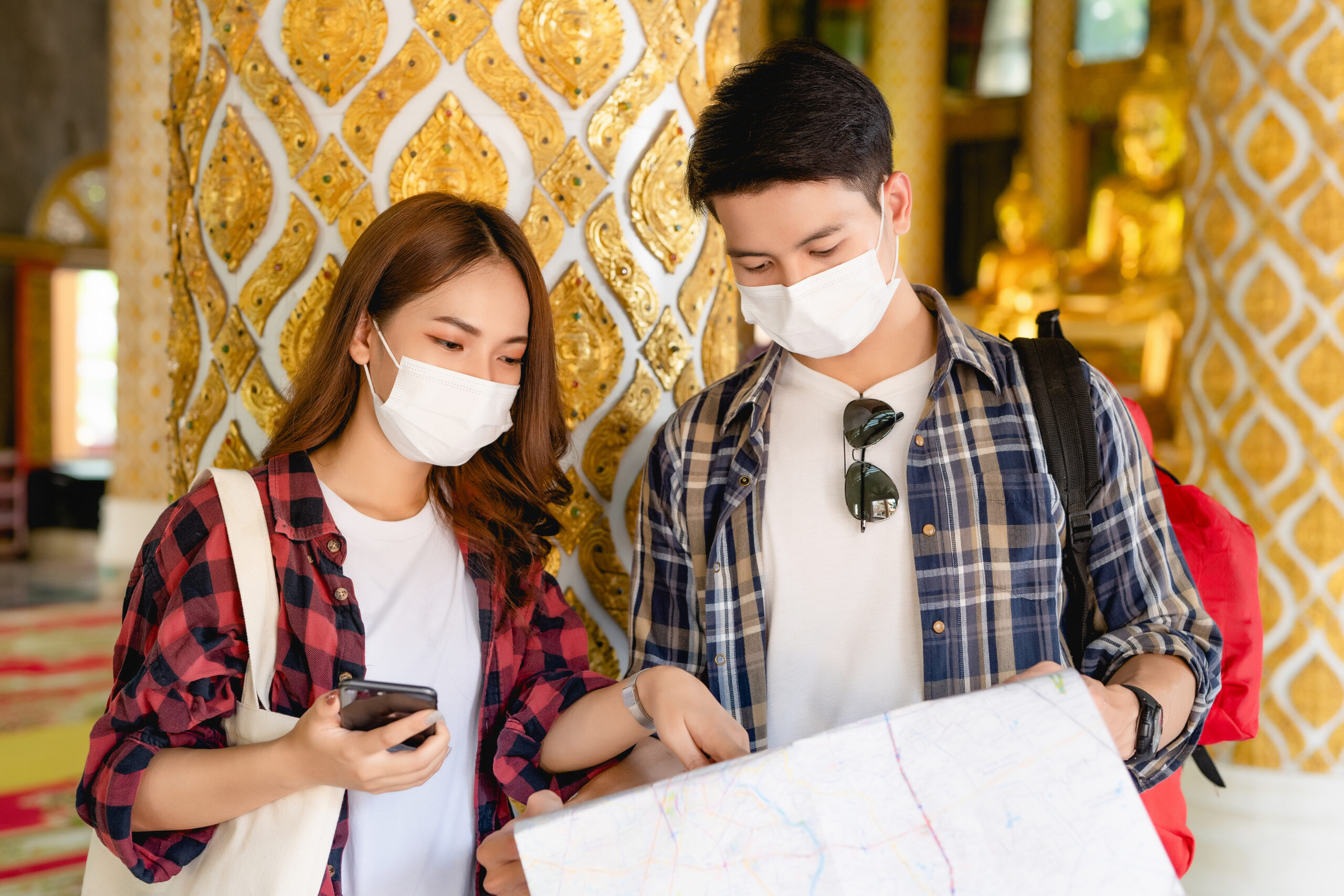 Asian couple tourist backpackers standing in beautiful Thai temple, pretty woman holding smartphone and handsome man check in paper map during travel on vacations